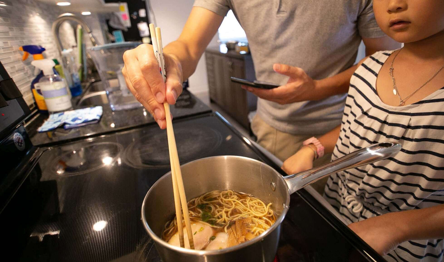 A parent cooking ramen with chopsticks while a child watches, highlighting a family cooking moment.