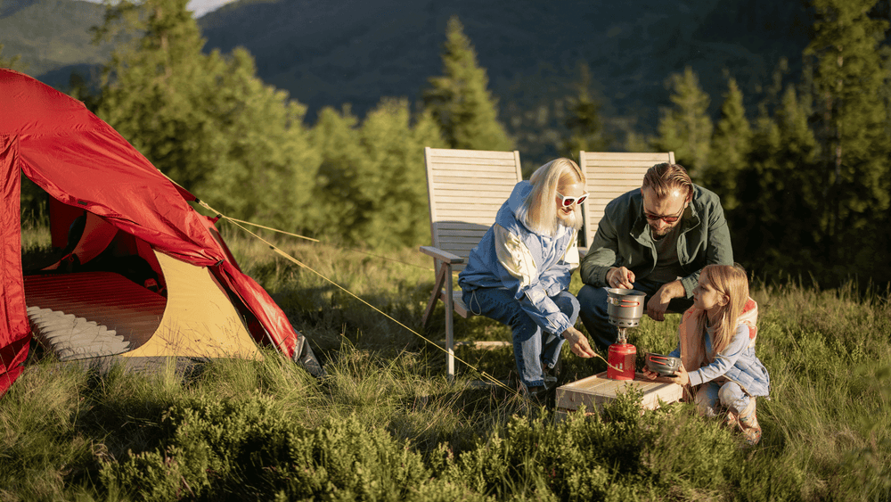 Family enjoying camping with a red tent and outdoor cooking equipment in a scenic forest setting.