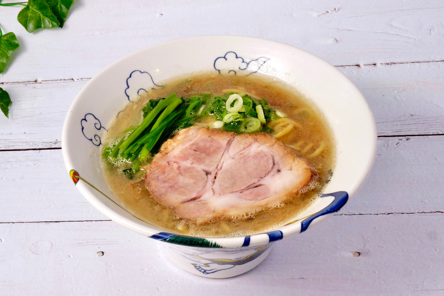 Bowl of Iekei ramen with rich broth, slices of pork, green onions, and leafy greens on a white wooden table