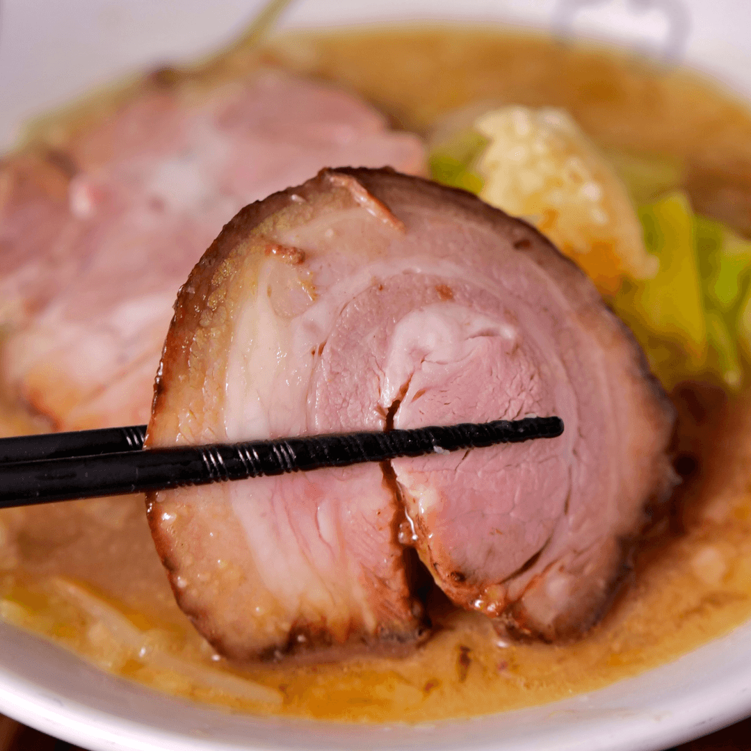 Sliced pork over a bowl of ramen with chopsticks holding a piece.